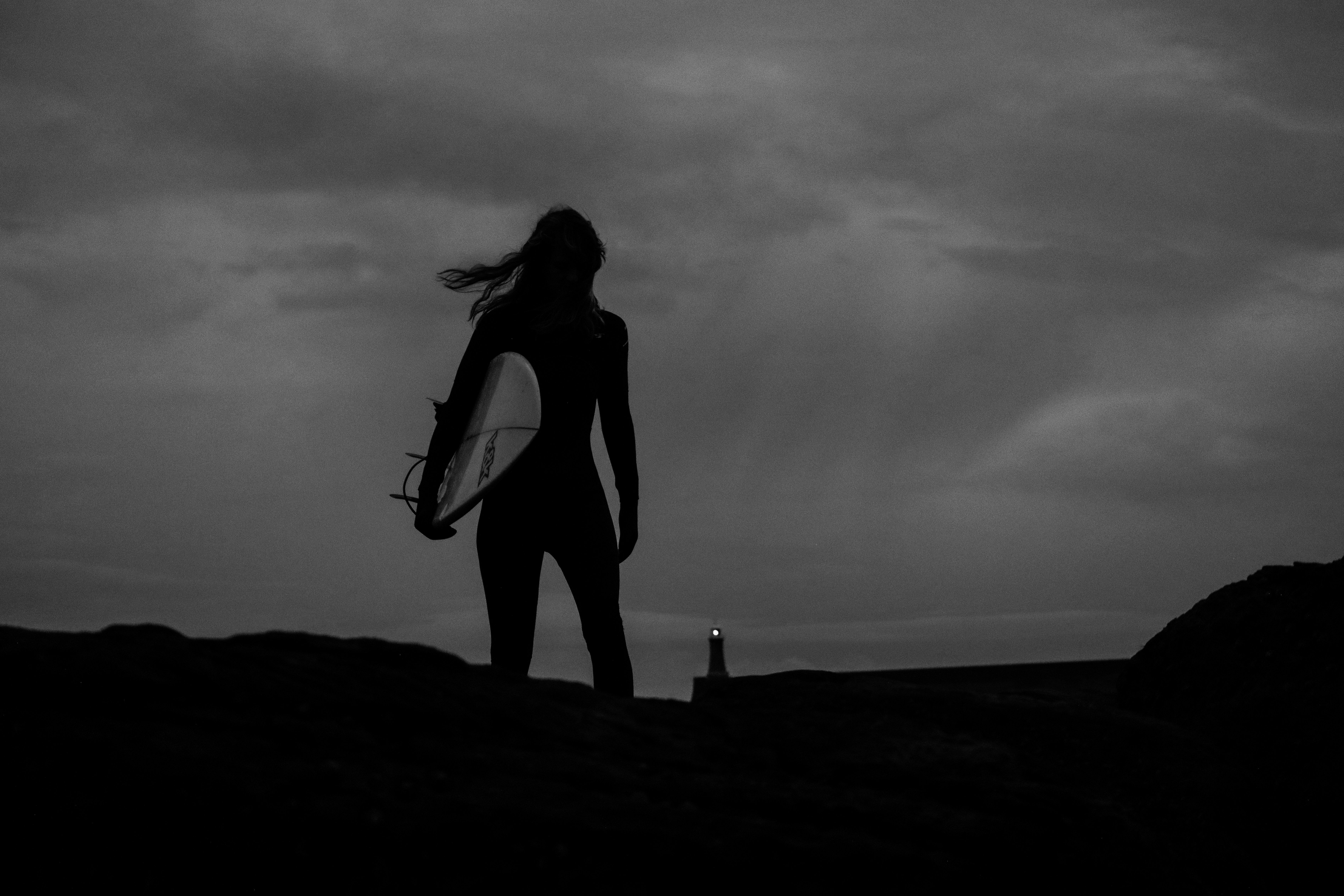 A female surfer is silhouetted against a cloudy sky in very low light, with a lighthouse on the horizon.